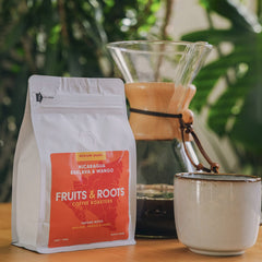 Fruits & Roots coffee bag with a coffee maker and cup on a wooden surface with greenery in the background