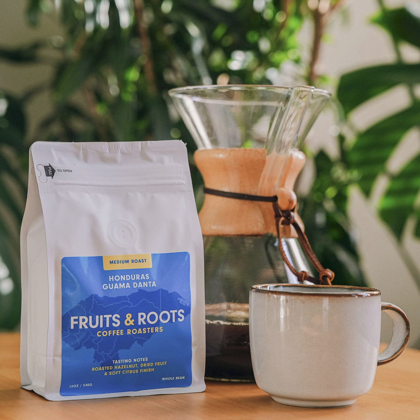 Fruits & Roots coffee bag with a cup and coffee maker on a wooden surface with plants in the background