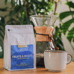 Fruits & Roots coffee bag with a cup and coffee maker on a wooden surface with plants in the background