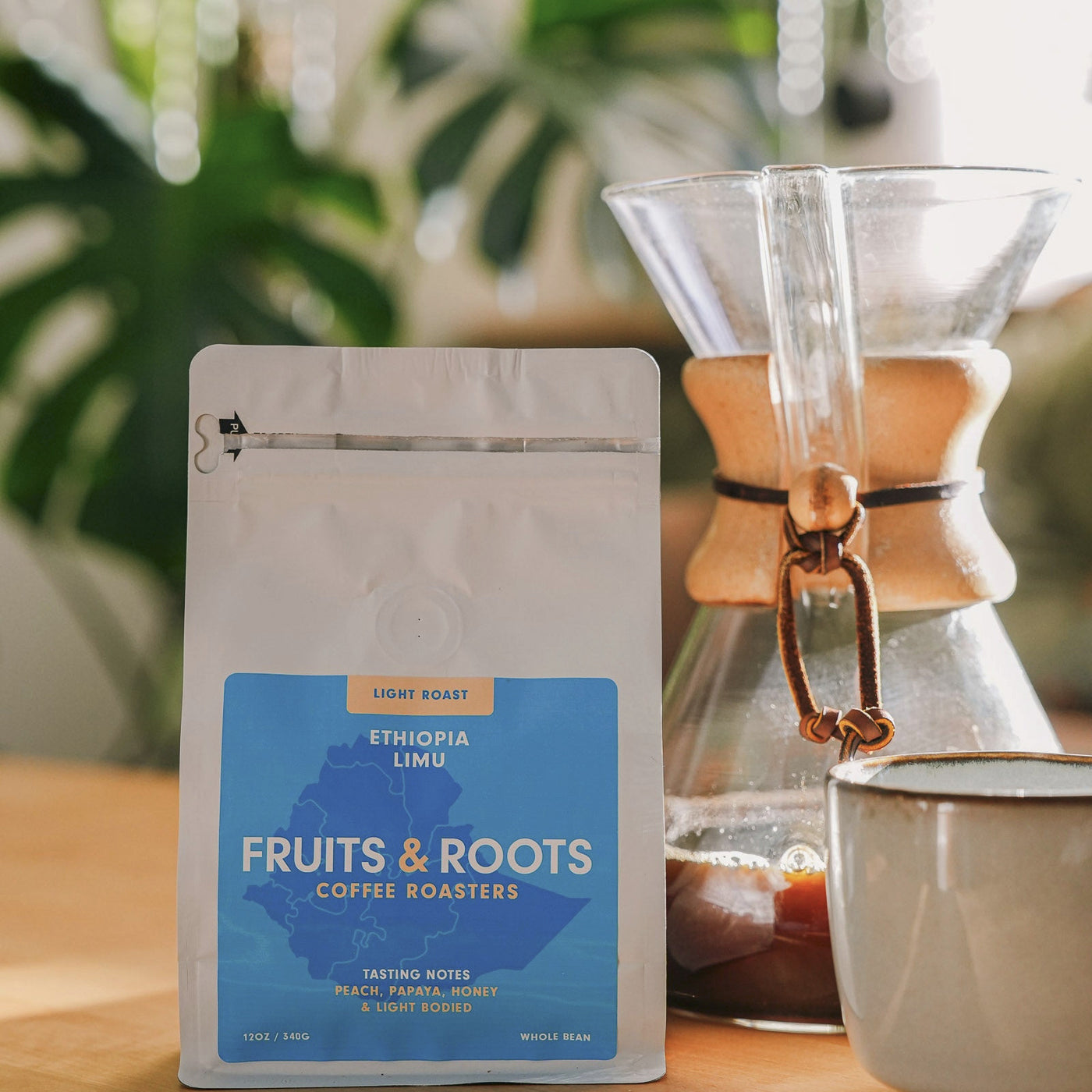 Fruits & Roots coffee package with a Chemex and mug on a wooden surface with plants in the background